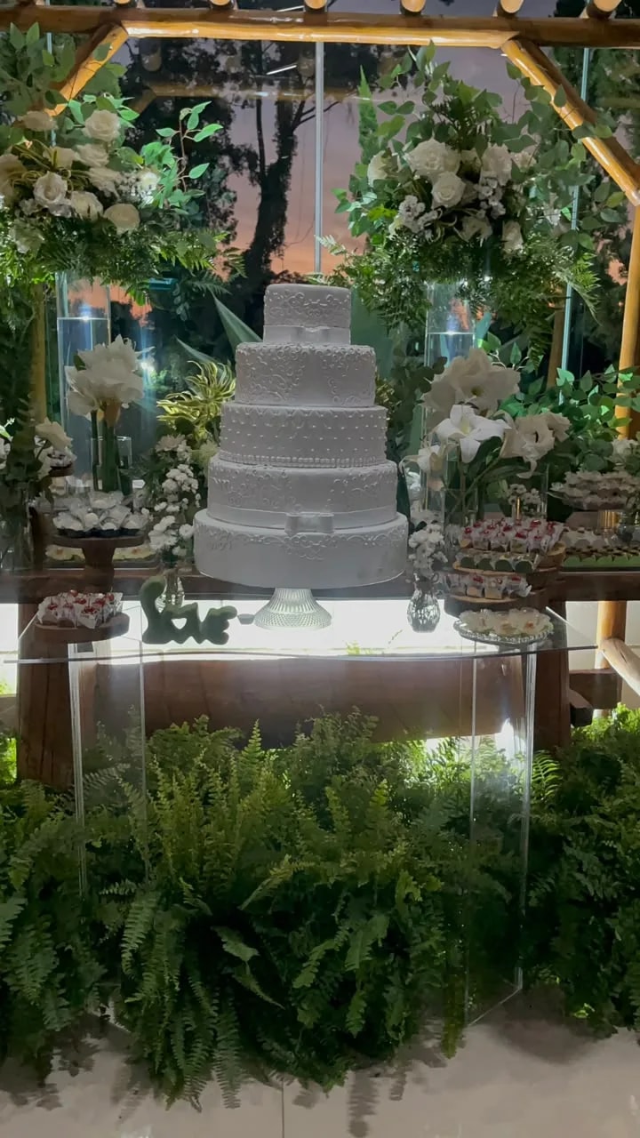 Mesa de bolo branco com flores brancas e samambaias sob o pergolado