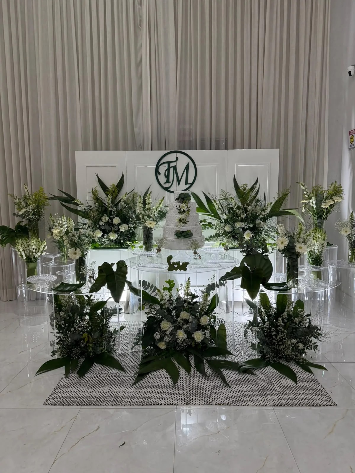 Mesa de bolo com folhagens e flores brancas diante de painel com monograma